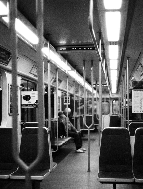 A person sits alone on a subway car with empty seats and bright overhead lights.