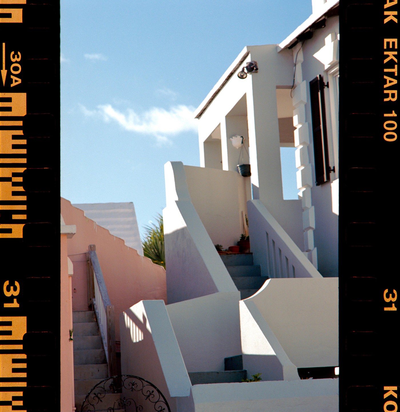 A white building with multiple staircases and a pink building are visible under a clear blue sky.