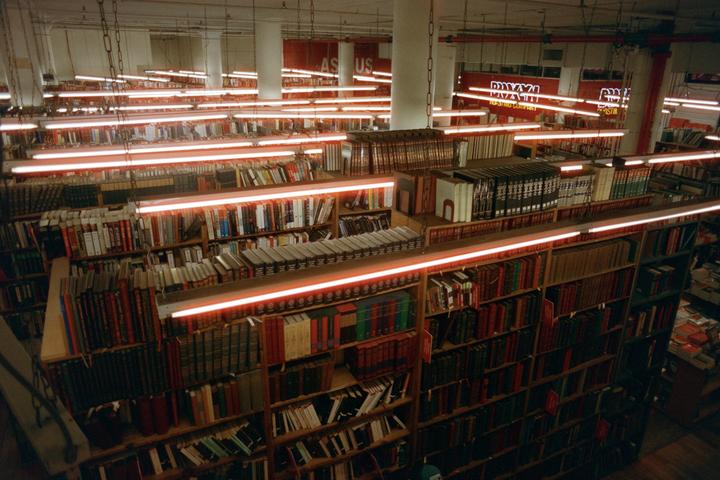 A high-angle view shows rows of bookshelves illuminated by long, glowing neon lights in a bookstore.