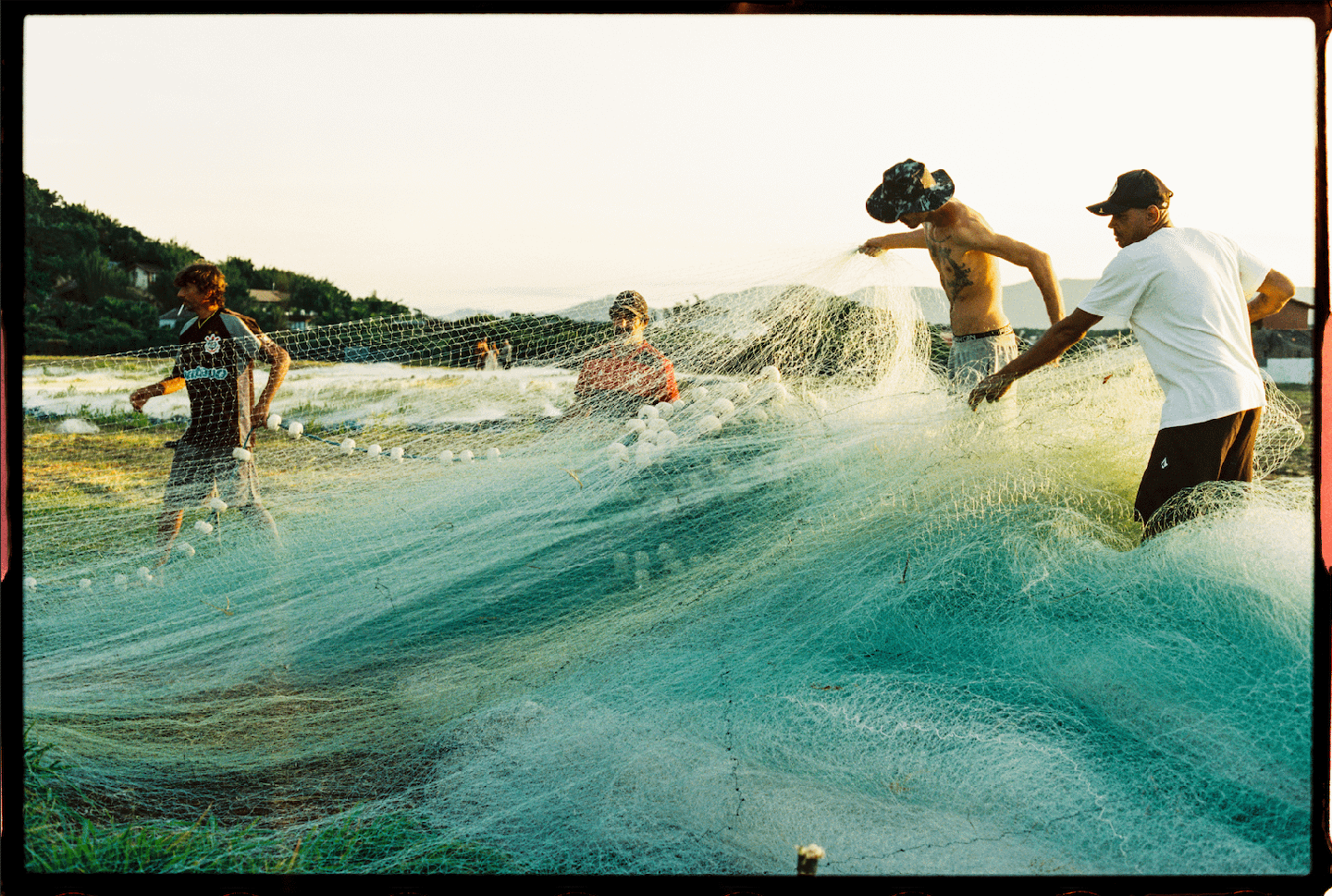 Four men are pulling a large fishing net from the water onto a grassy shore.