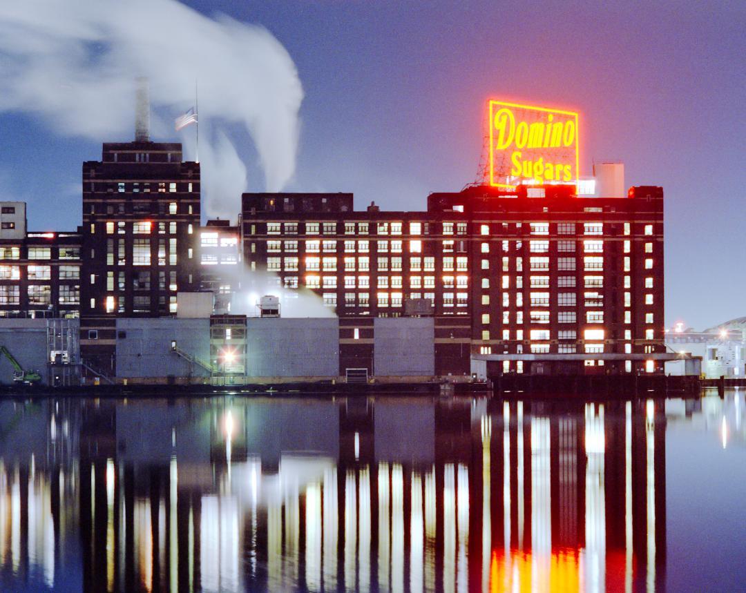 A large building with a neon sign for "Domino Sugars" is reflected in the water at night.