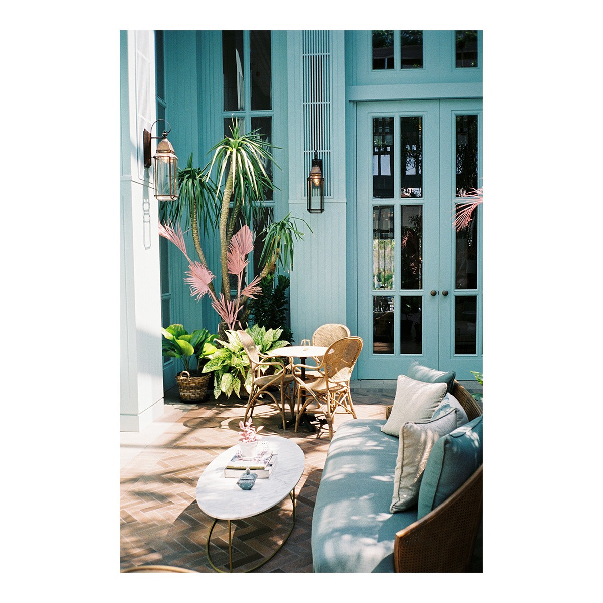 A patio with wicker furniture and potted plants sits in front of a light blue building with large windows and doors.