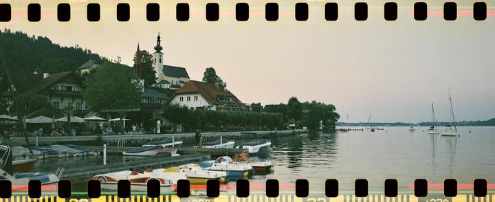 A small town with a church and houses is nestled on a hillside next to a lake with boats docked at a pier and sailboats in the water.