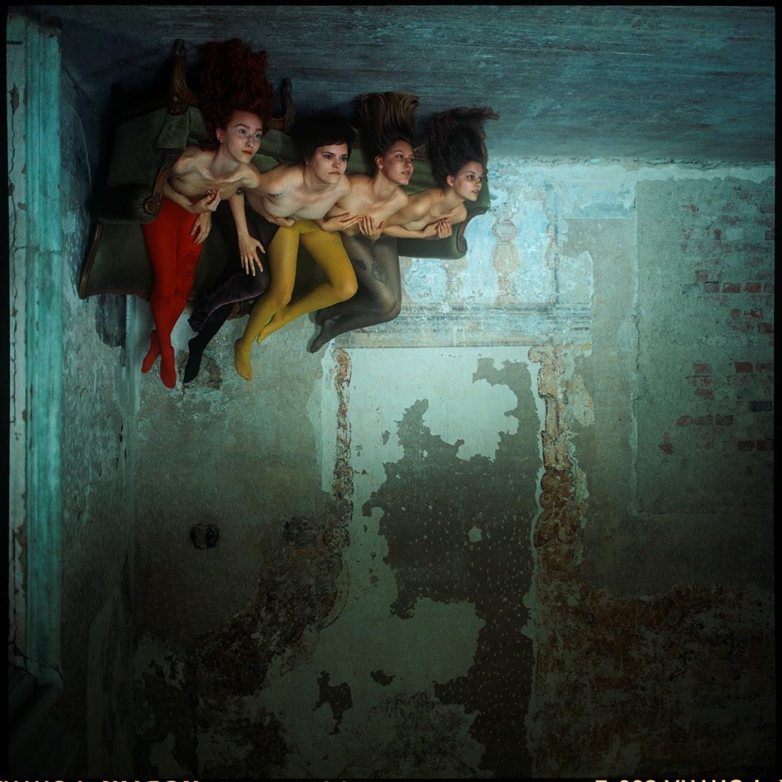 Four women with long hair are lying on a couch, wearing colorful tights, in a room with peeling paint on the walls.