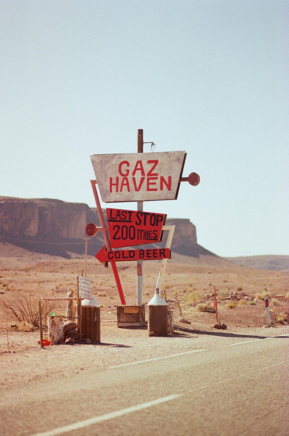 A weathered sign for Gaz Haven, advertising "Last Stop 200 Miles" and "Cold Beer," stands beside a desert road with mountains in the background.
