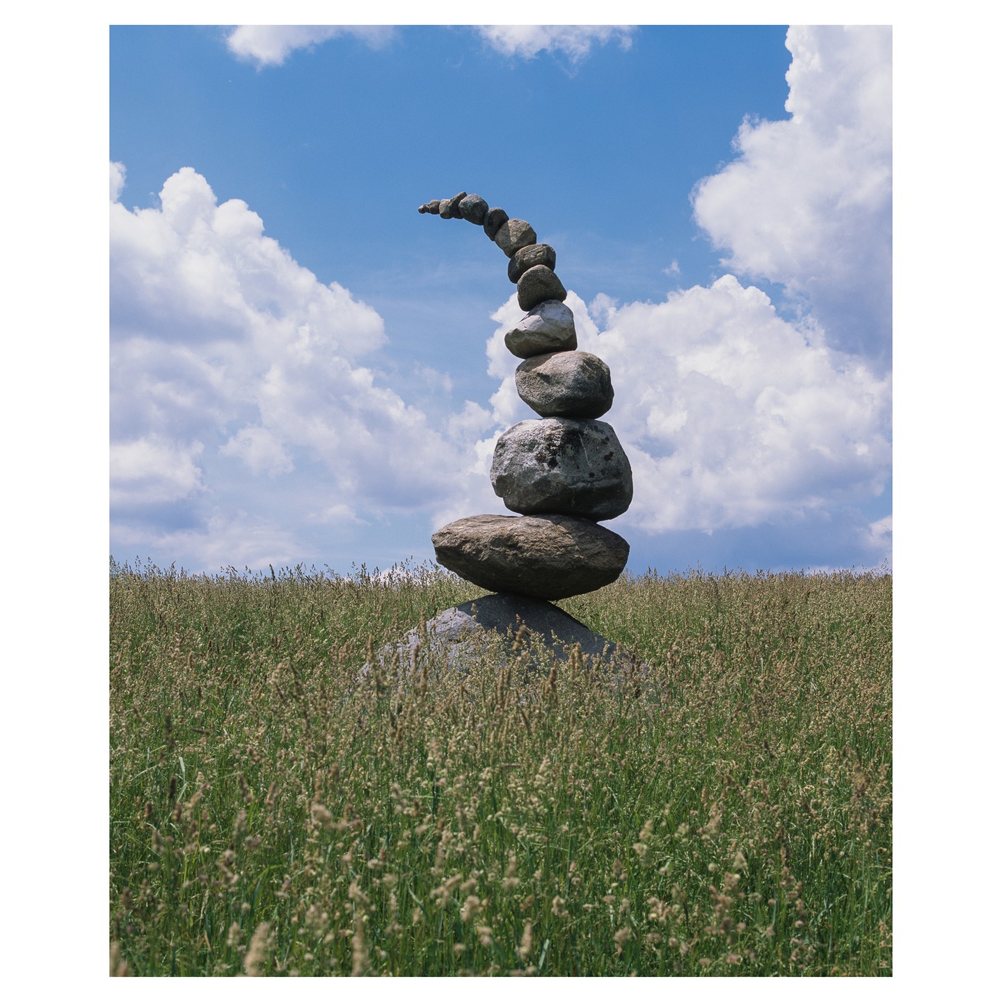 A tall stack of balanced rocks stands in a field of tall grass under a blue sky with white clouds.