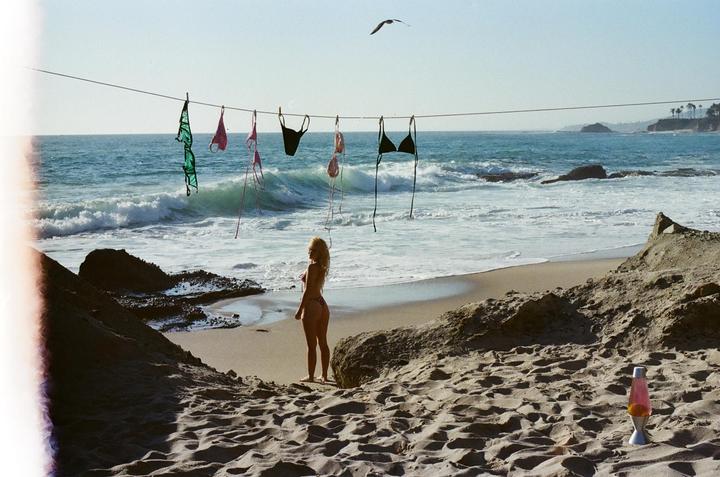 A woman stands on a sandy beach with several bikini tops hanging on a line above the ocean.