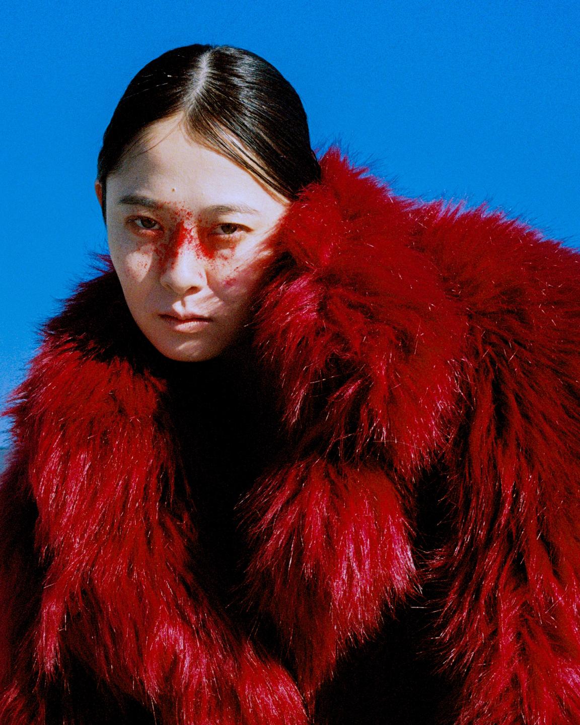 A woman with red makeup on her face is wearing a red fur coat against a blue background.