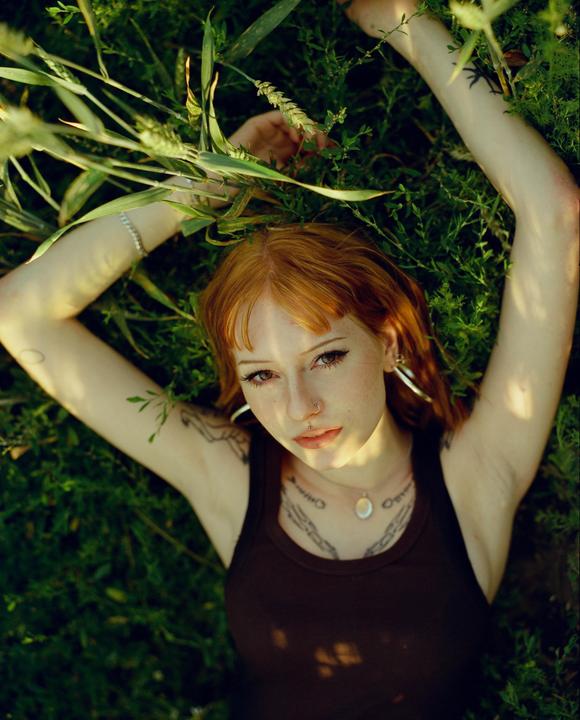 A woman with red hair and tattoos lies in green grass with her arms raised above her head.