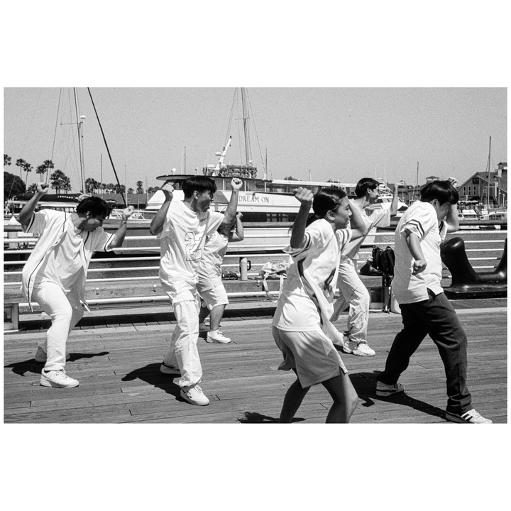 A group of children are dancing on a wooden pier with boats and palm trees in the background.