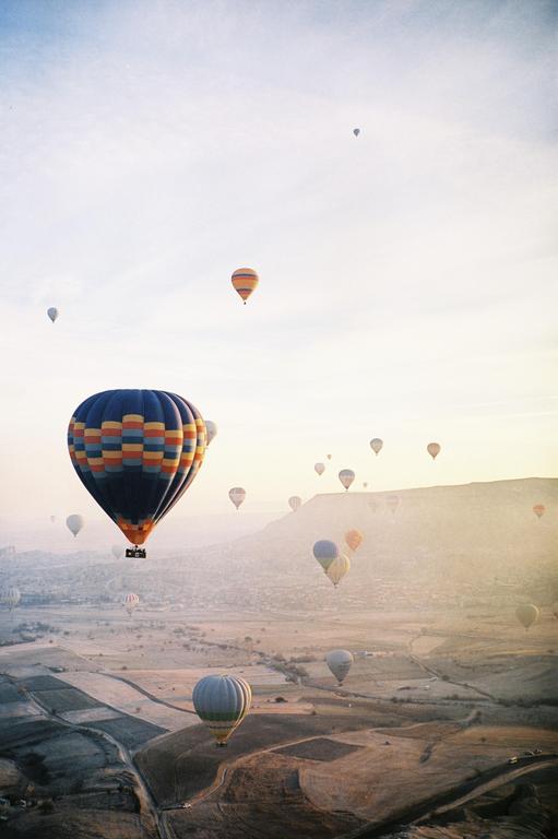Many hot air balloons float above a hazy, mountainous landscape at dawn.