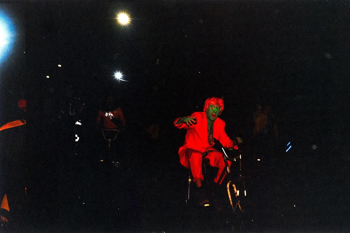 A person with green face paint and red hair, wearing a red suit, rides a bicycle at night.