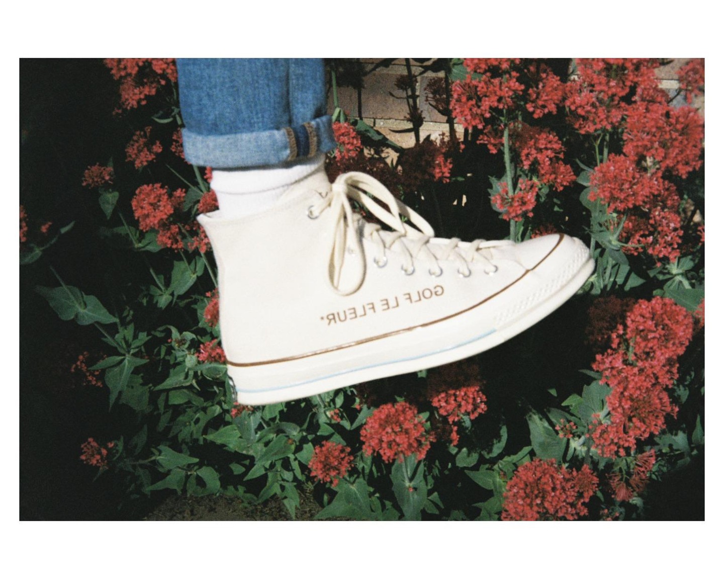 A white high-top sneaker with blue jeans is surrounded by red flowers and green foliage.