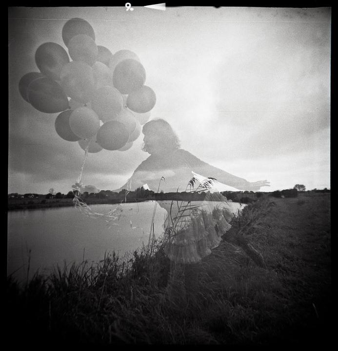 A double exposure shows a woman holding balloons superimposed over a landscape with a body of water and tall grass.