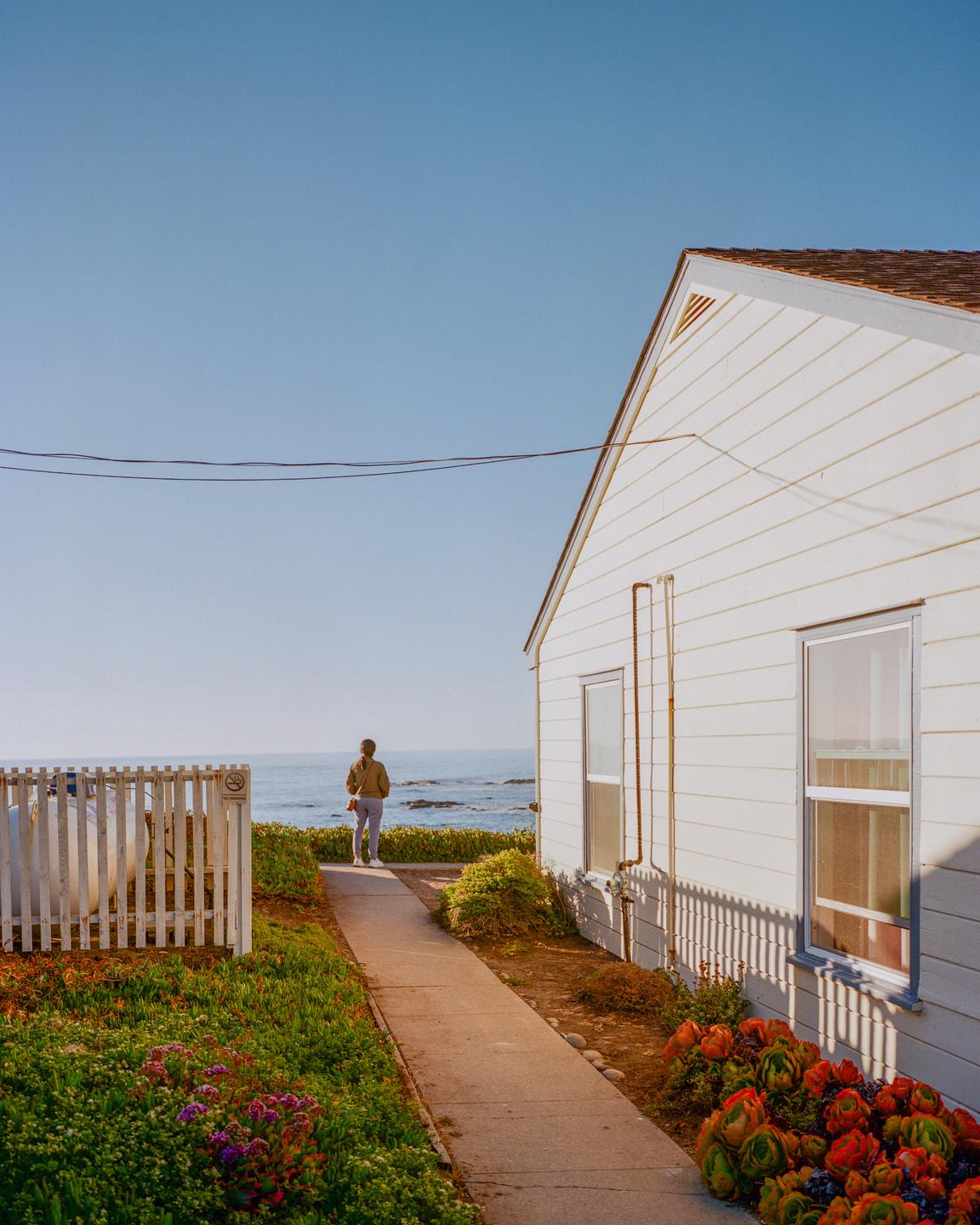 A person walks along a path towards the ocean next to a white house with a picket fence.
