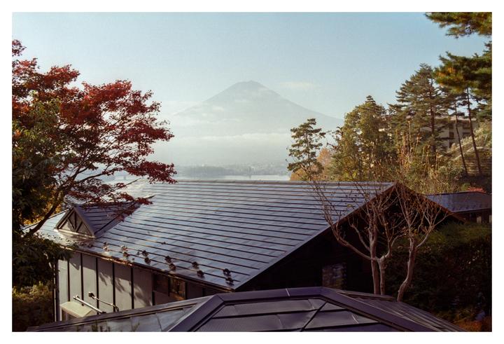 A mountain rises in the distance behind a lake and several buildings with dark roofs, surrounded by trees with autumn foliage.