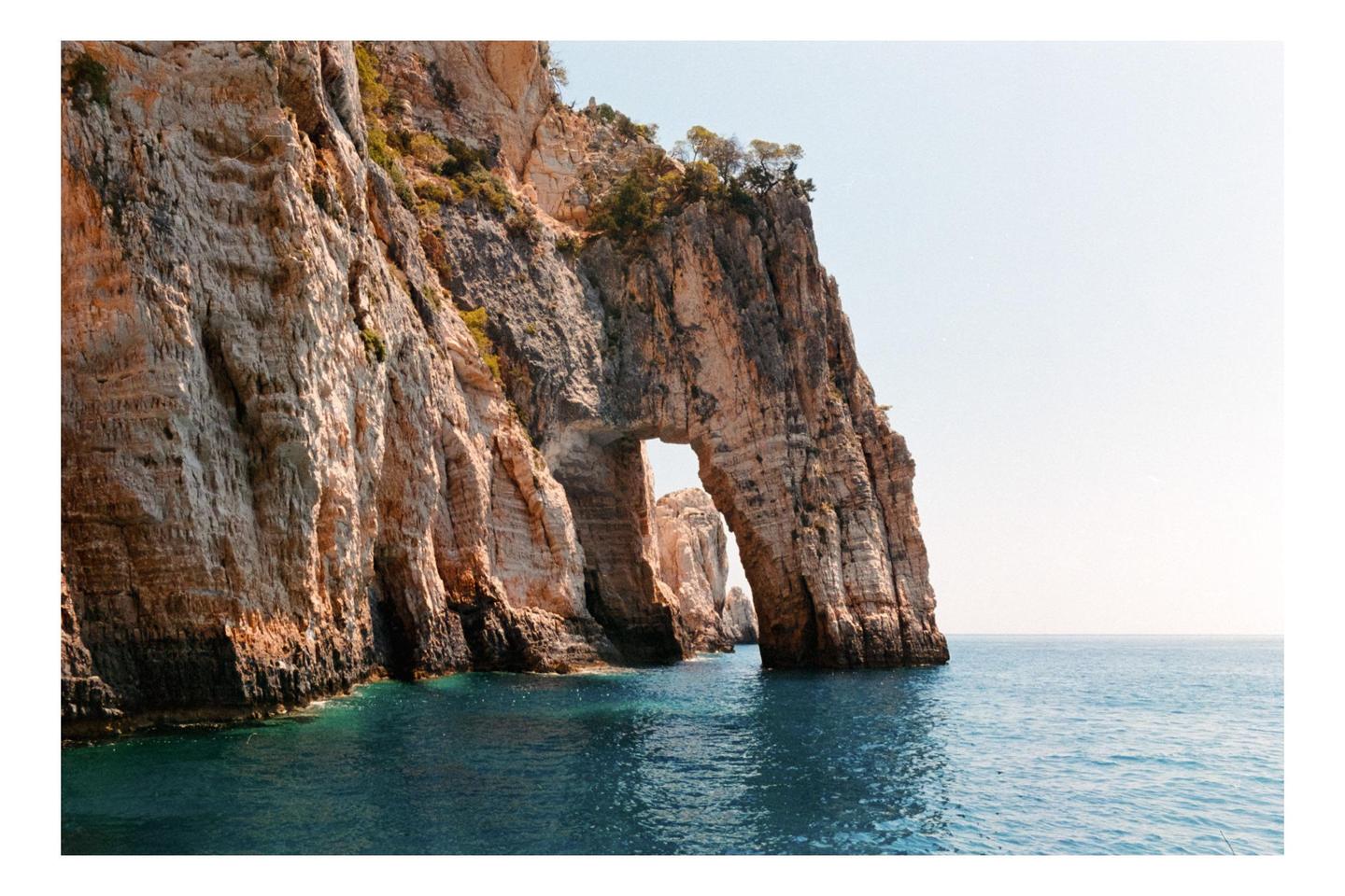 A large rock formation with an archway stands in the blue ocean under a clear sky.