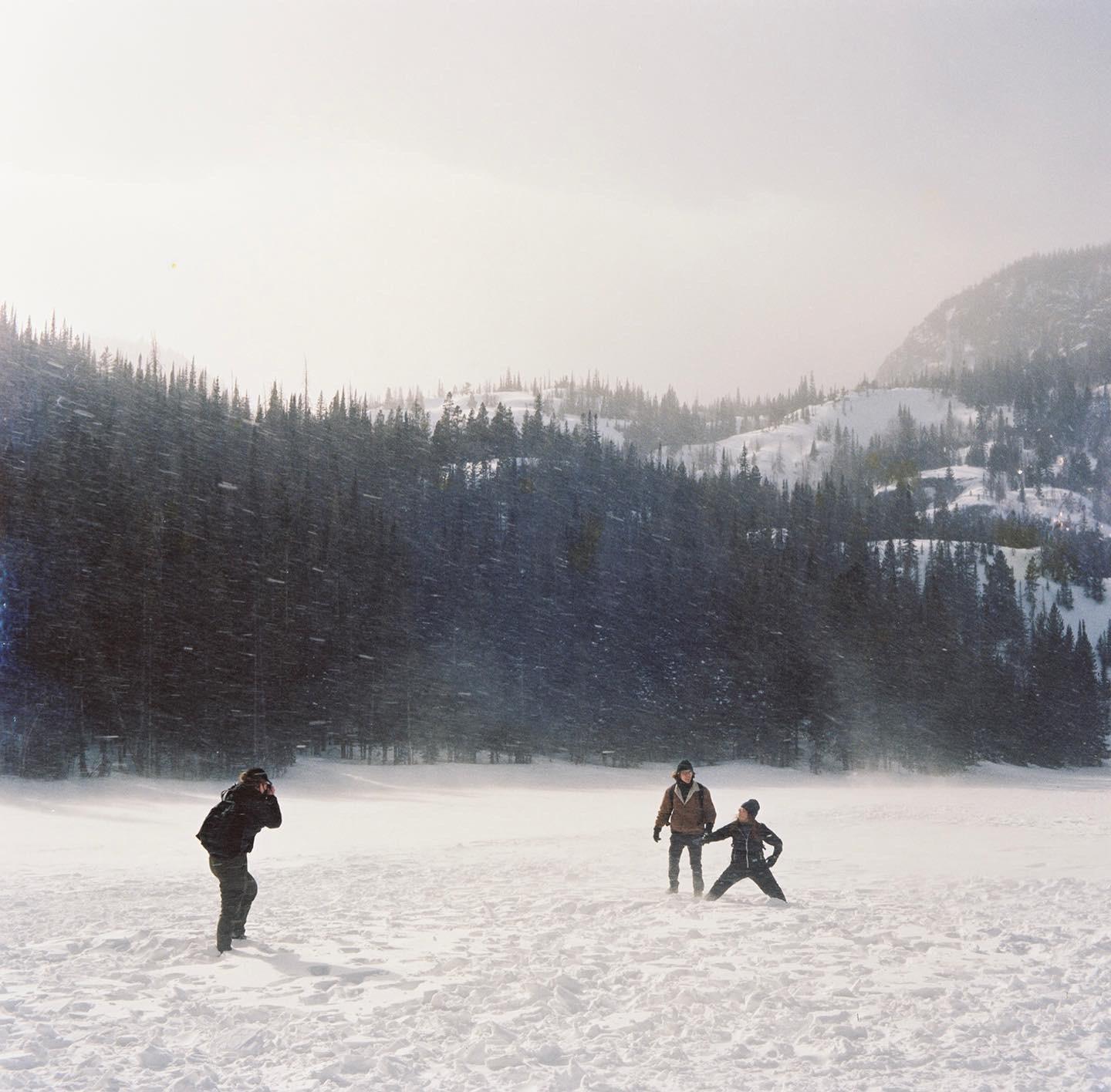 Three people are in a snowy field with a forest and mountains in the background.
