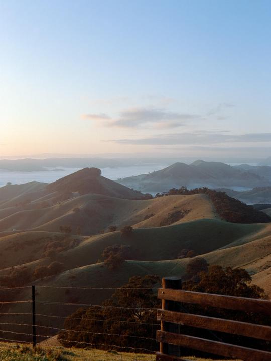 A wooden fence and barbed wire fence are in the foreground of rolling hills covered in fog.