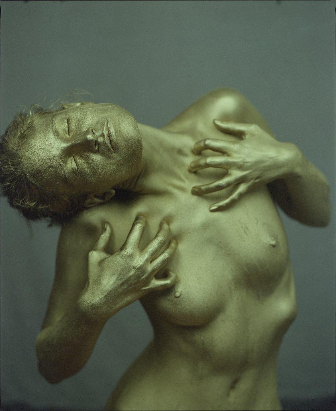 A woman with her eyes closed and her body covered in gold paint is shown from the chest up.