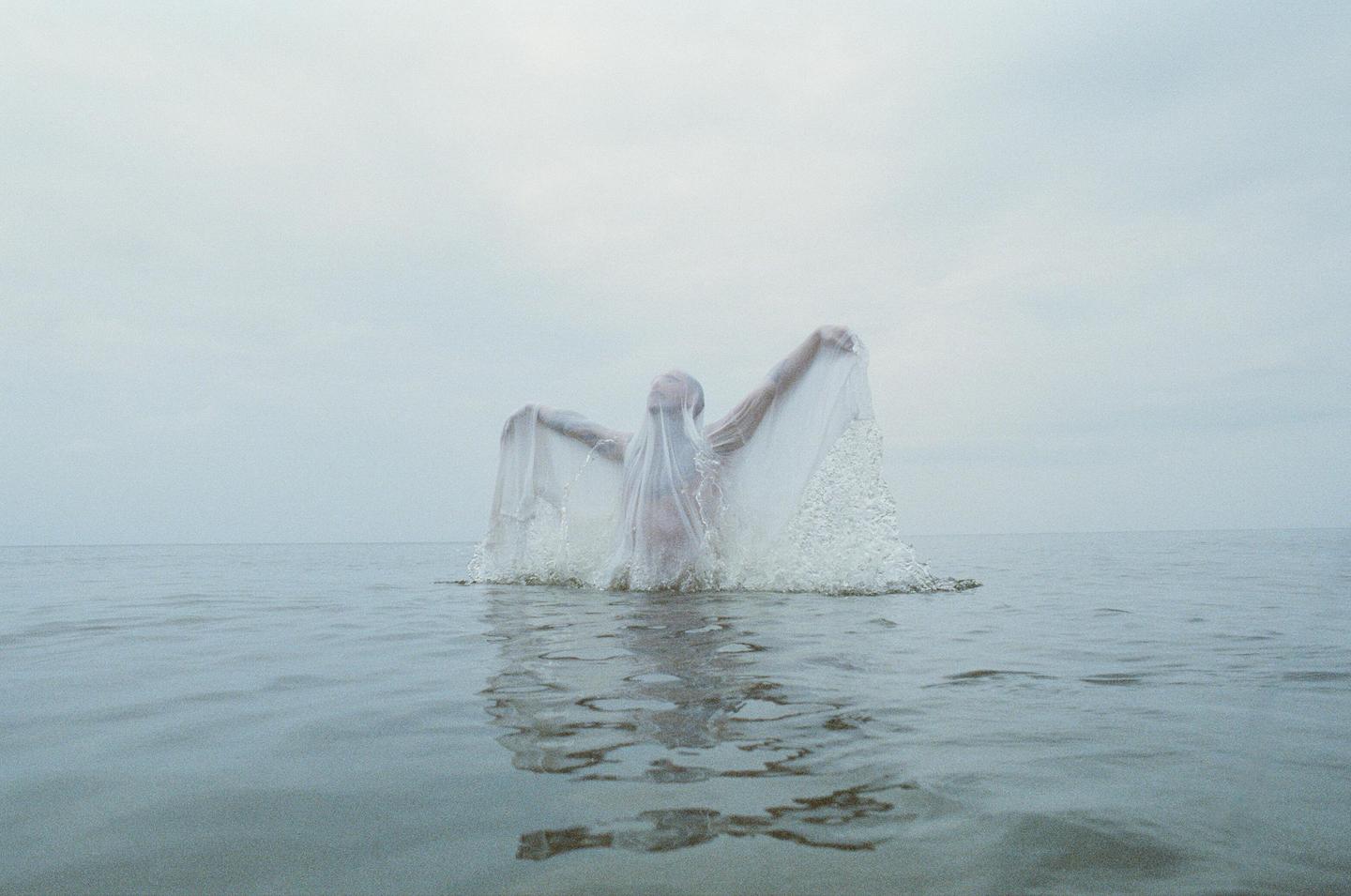 A person emerges from the water covered in a white translucent fabric that creates a splash.