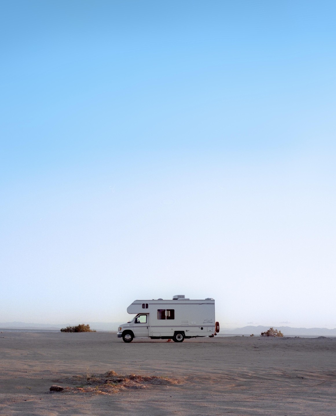 A white recreational vehicle is parked on a vast, flat, sandy plain under a clear sky.