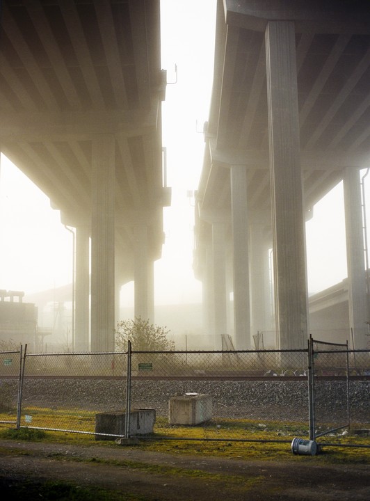 A foggy morning shows the concrete pillars of an overpass towering above a chain link fence and railroad tracks.
