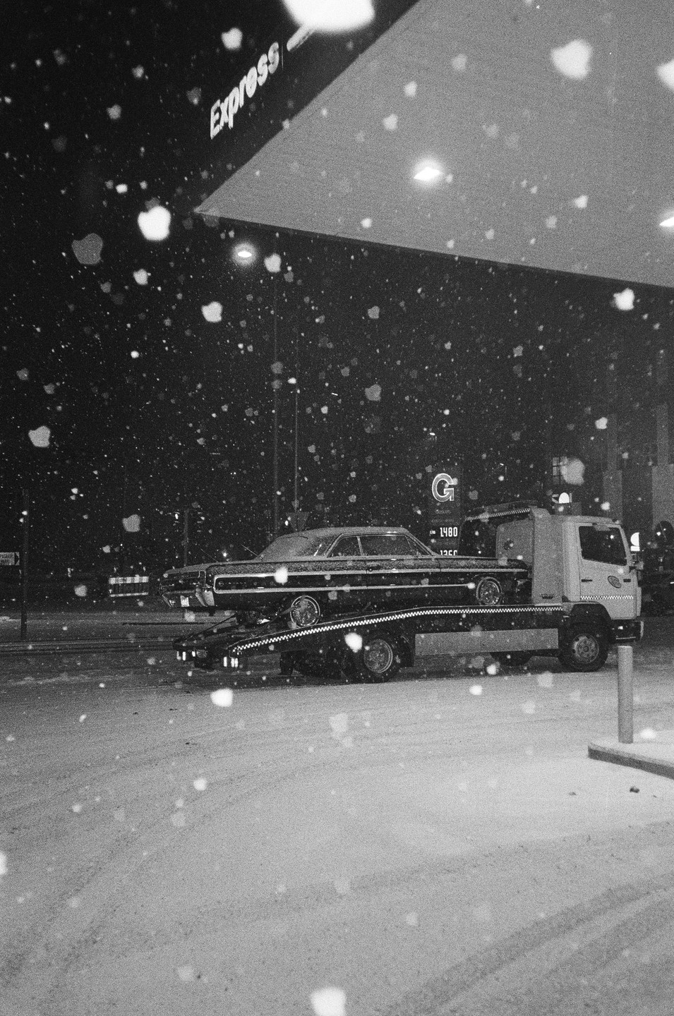 A tow truck carries a car on a snowy night at a gas station.