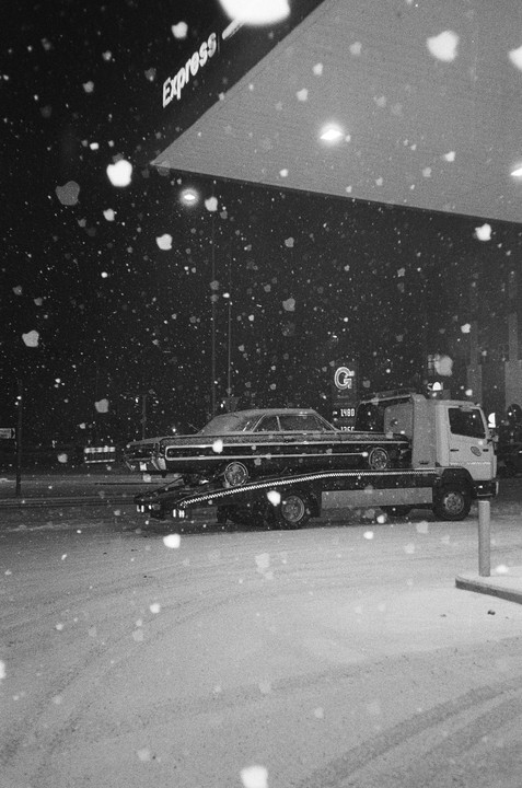 A tow truck carries a car on a snowy night at a gas station.