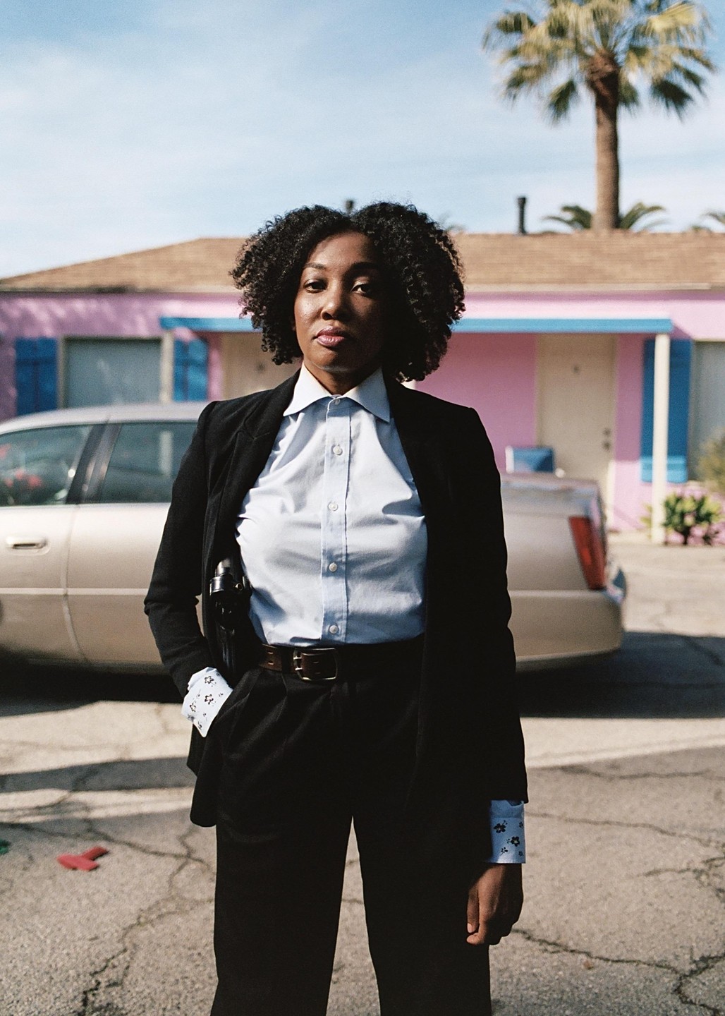 A woman in a suit with a holstered pistol stands in front of a pink building and a car.