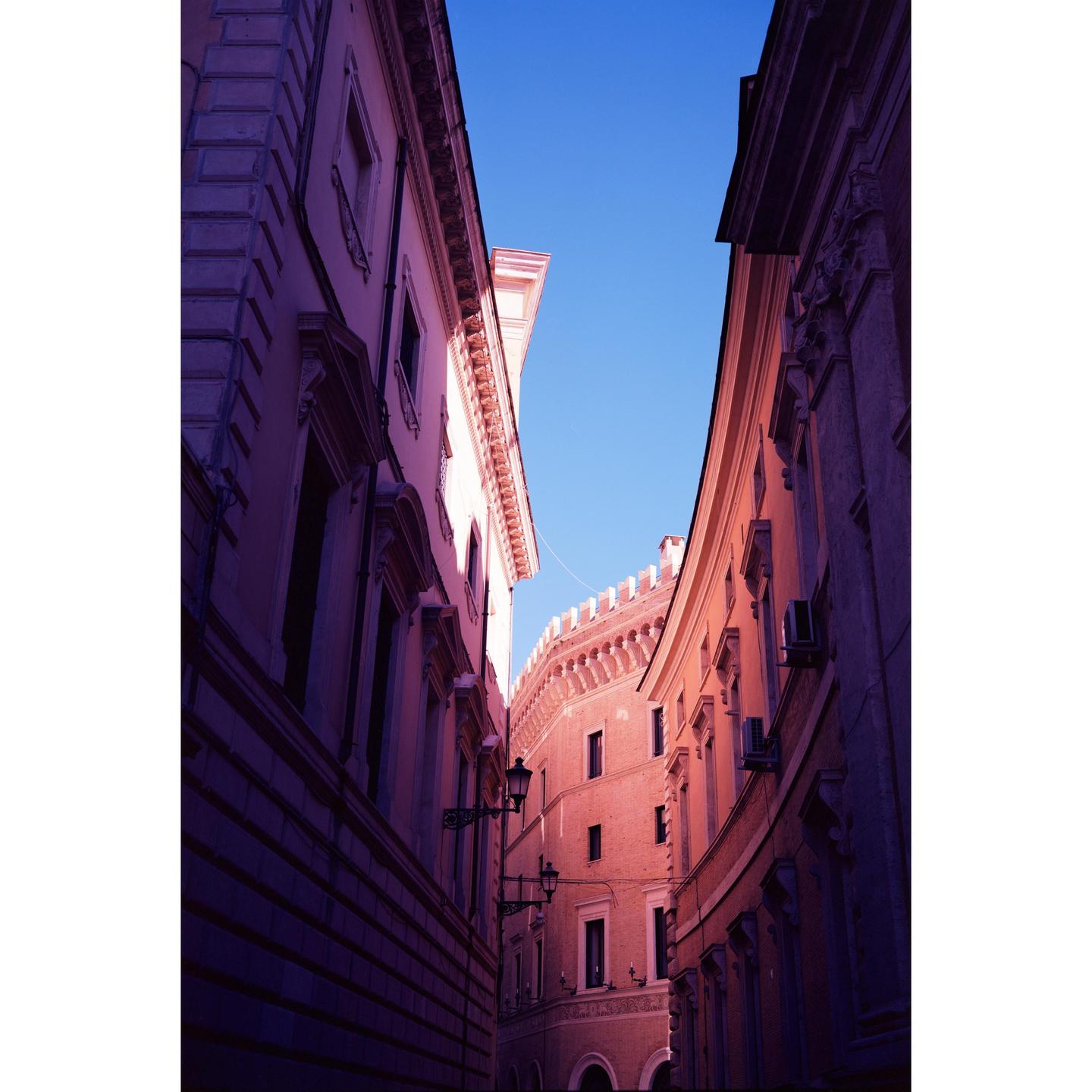 A narrow street in Rome is lined with tall buildings under a clear blue sky.