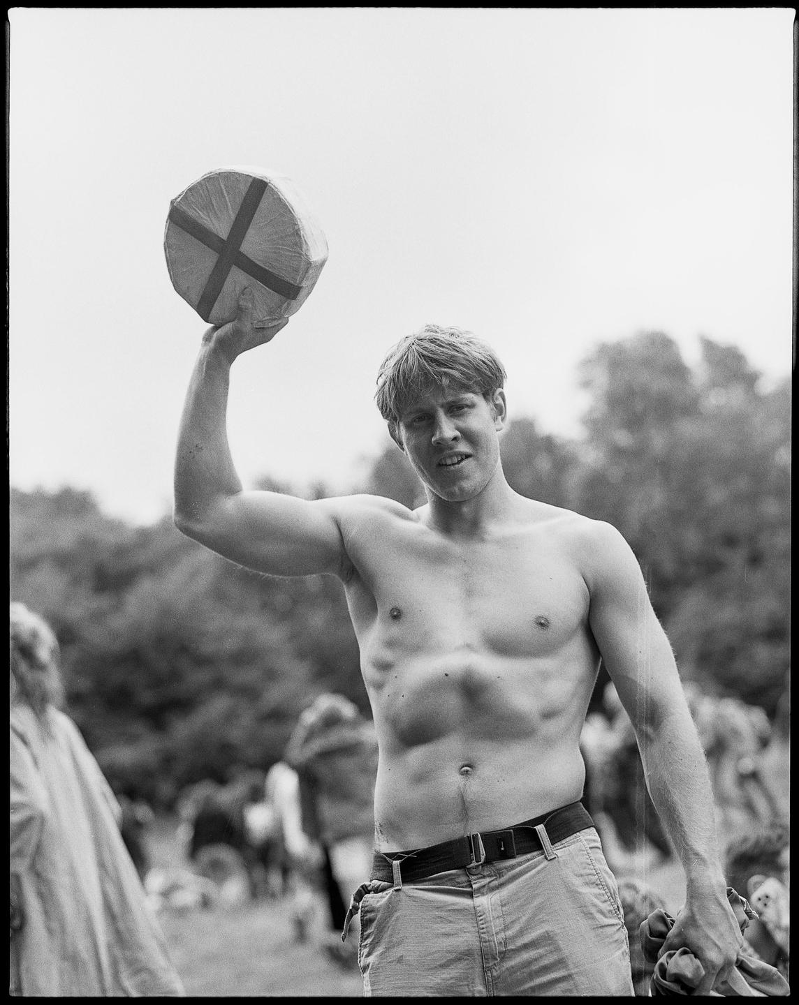 A shirtless man holds a cheese wheel above his head at an outdoor event.