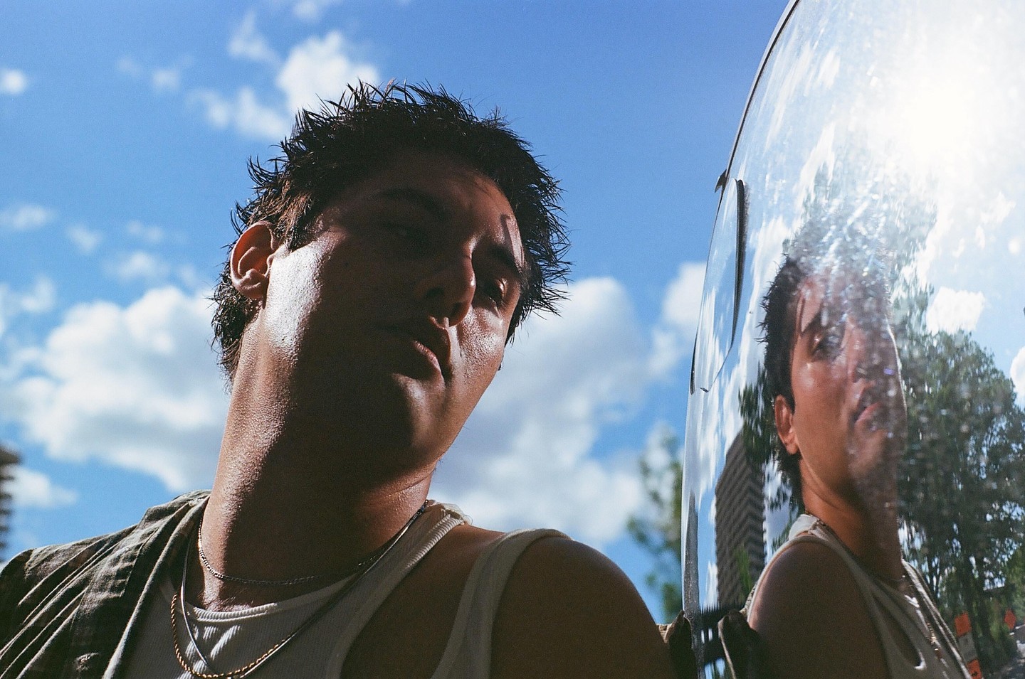 A man with wet hair and a tank top looks down at the camera, with his reflection visible in a shiny surface next to him.