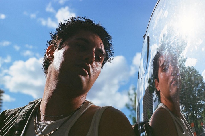 A man with wet hair and a tank top looks down at the camera, with his reflection visible in a shiny surface next to him.
