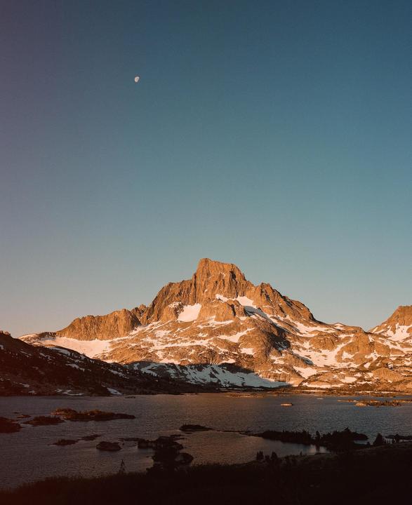 A moon is visible in the clear sky above a snow-capped mountain and a lake.