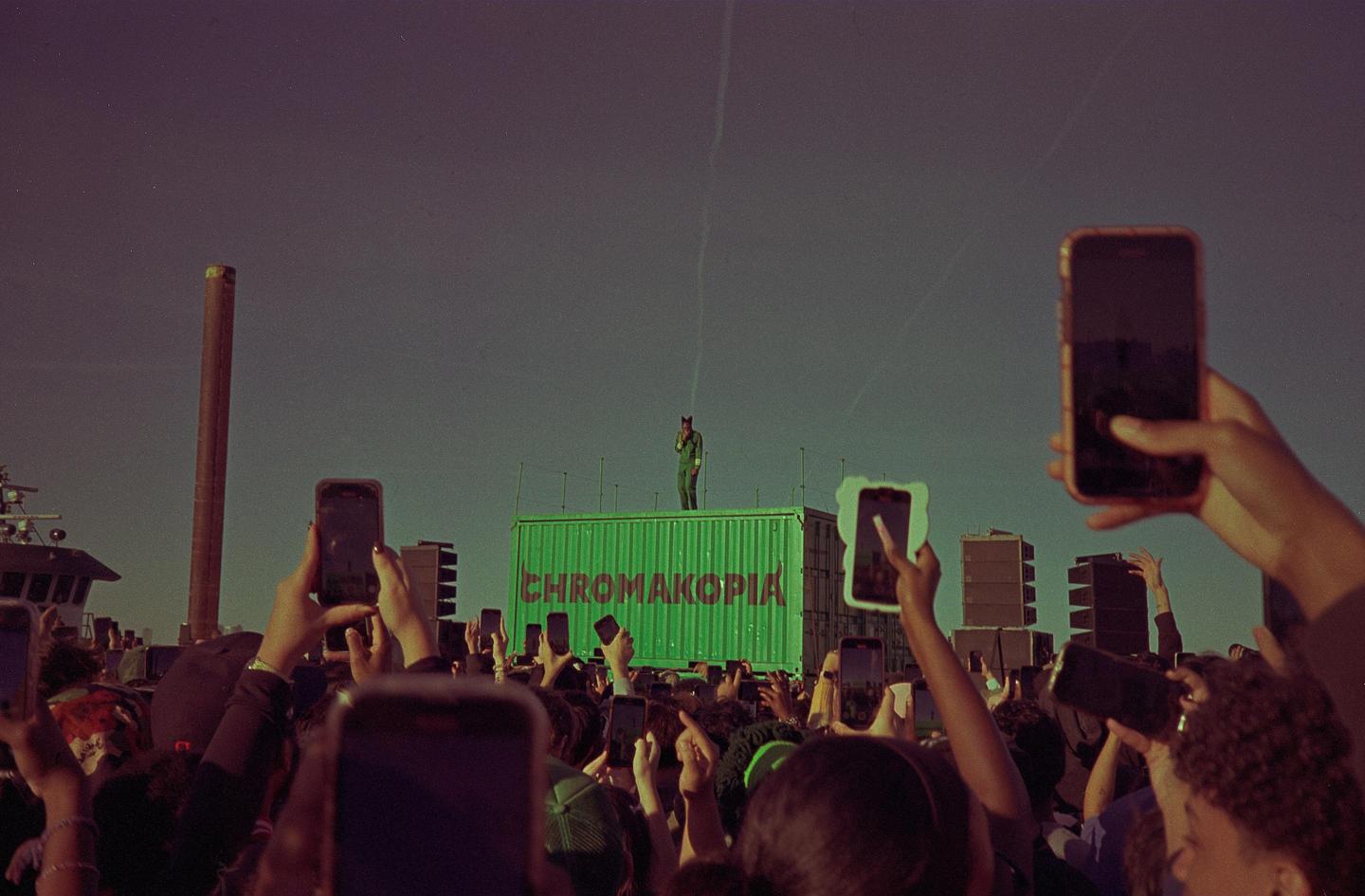 A person stands on a green container with "CHROMAKOPIA" written on it, performing for a crowd of people holding up phones.