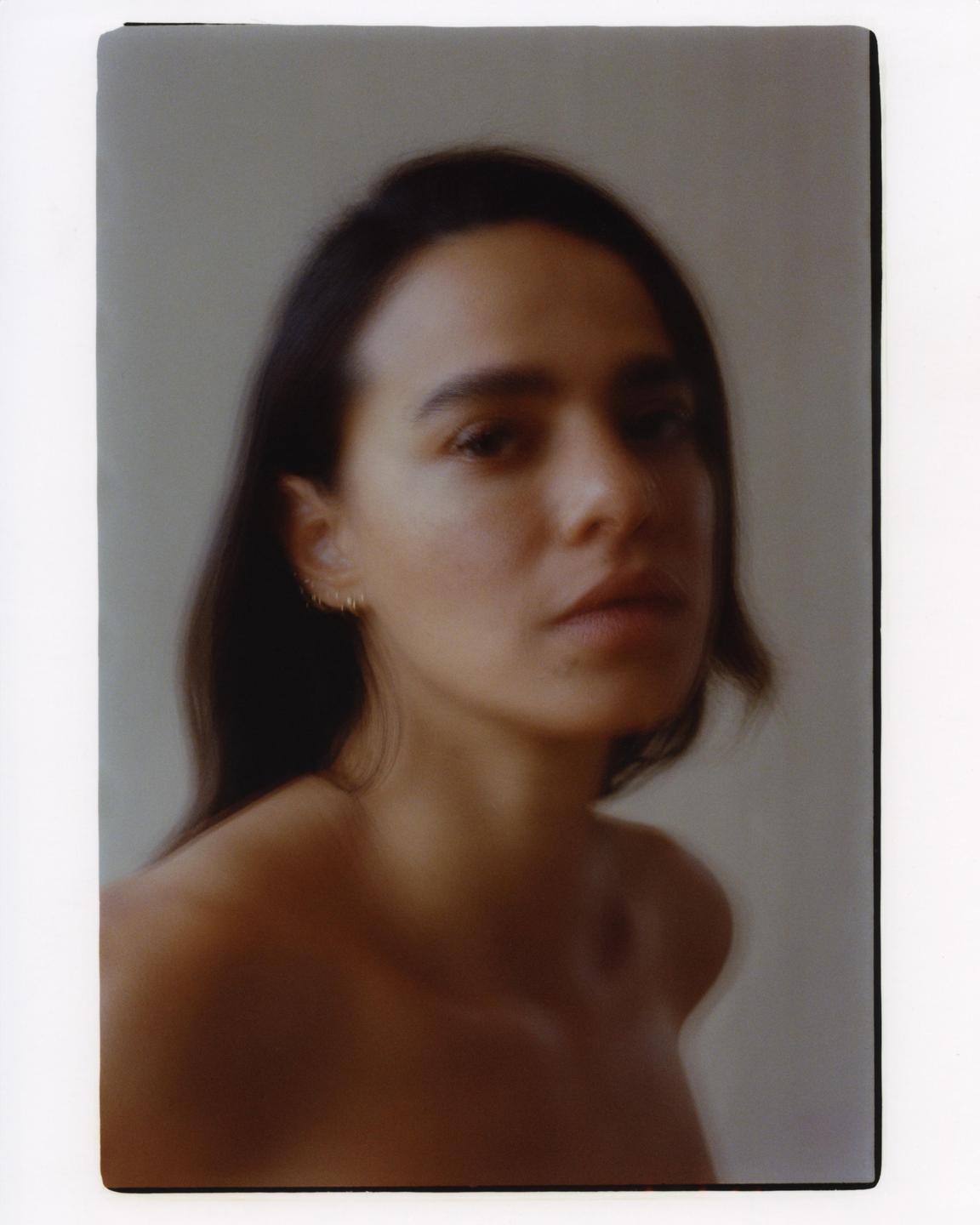 A woman with dark hair and bare shoulders looks directly at the viewer.
