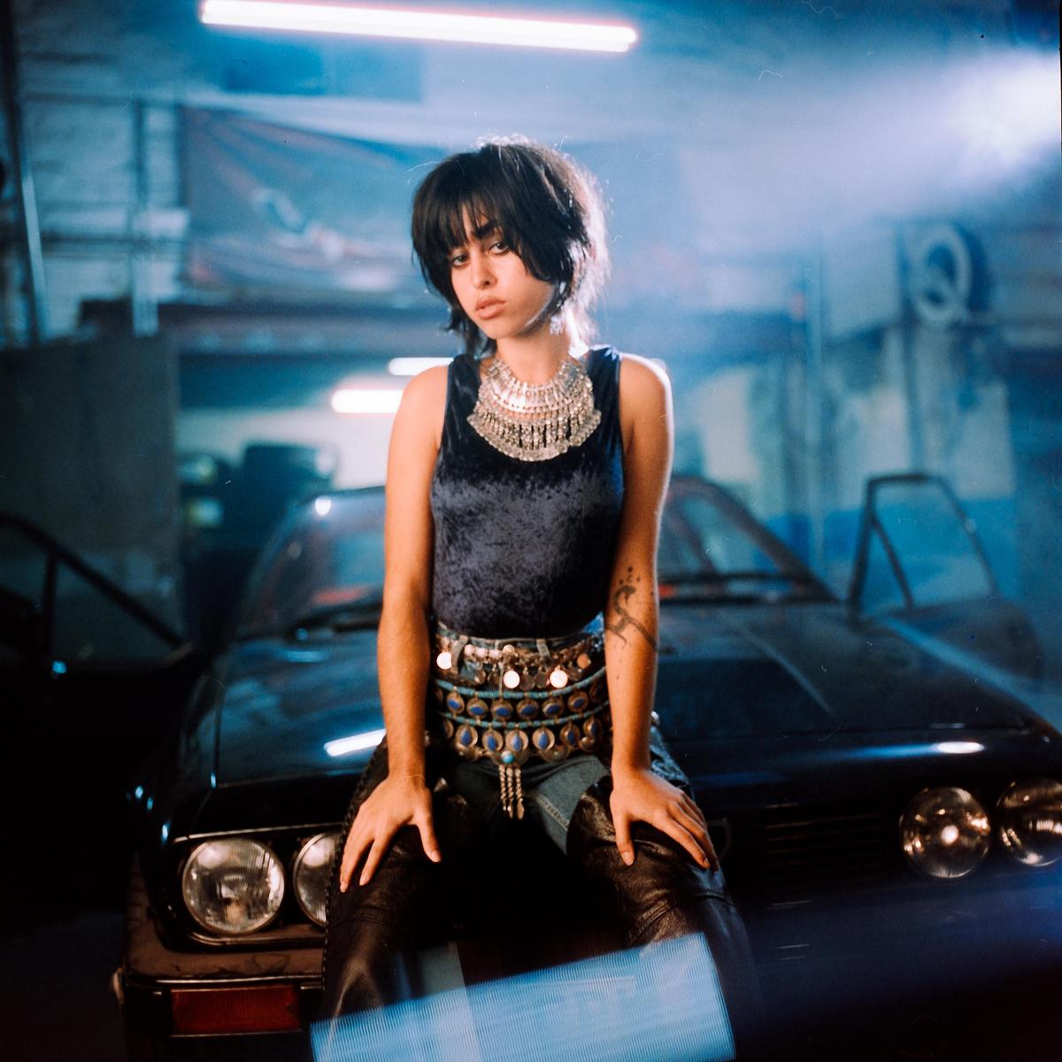 A woman with dark hair and a velvet top sits on the hood of a car in a garage.