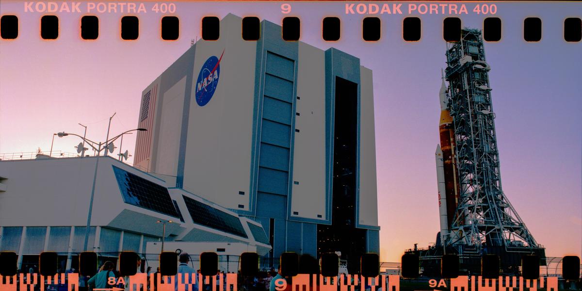 A large rocket stands next to a tall building with the NASA logo at sunset.