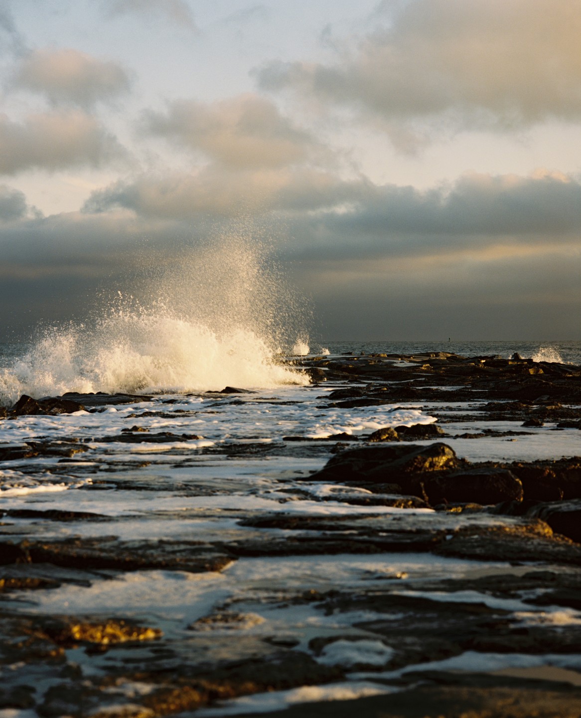 A wave crashes against a rocky shore under a cloudy sky.