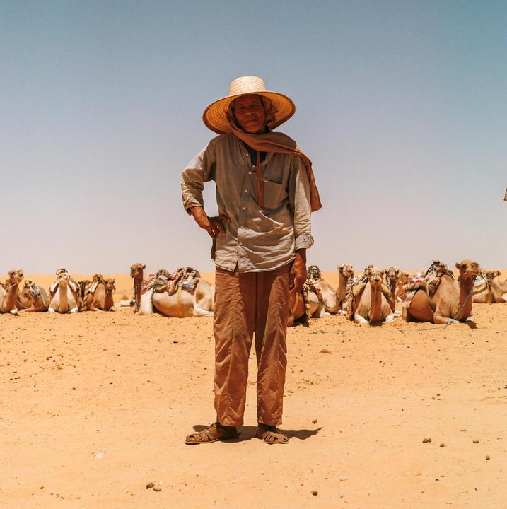 A man in a straw hat stands in the desert with many camels resting behind him.