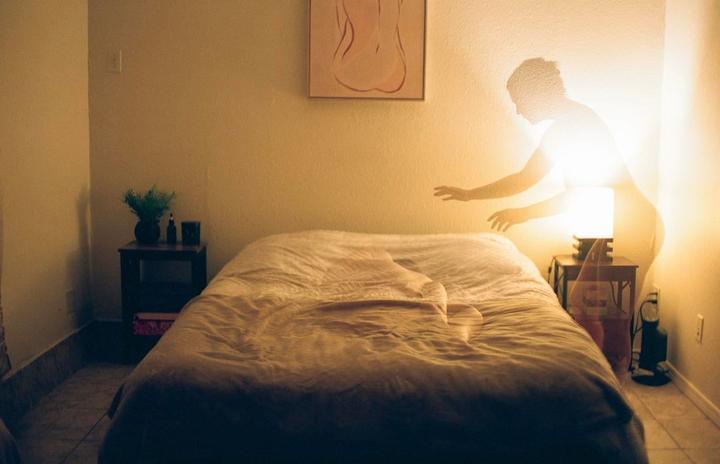 A double exposure shows a glowing figure reaching over a bed in a bedroom.