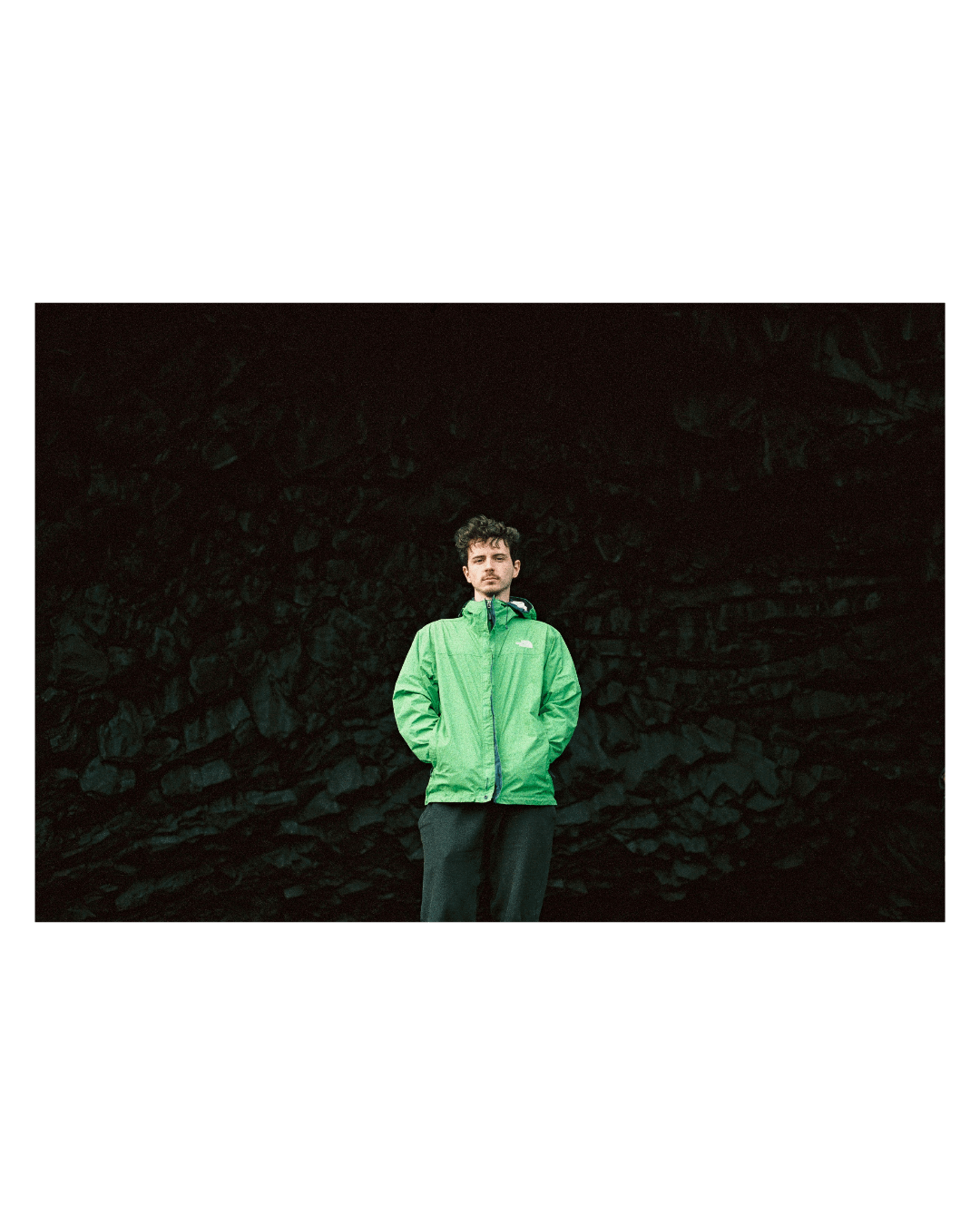 A man in a green jacket stands against a dark, rocky background.