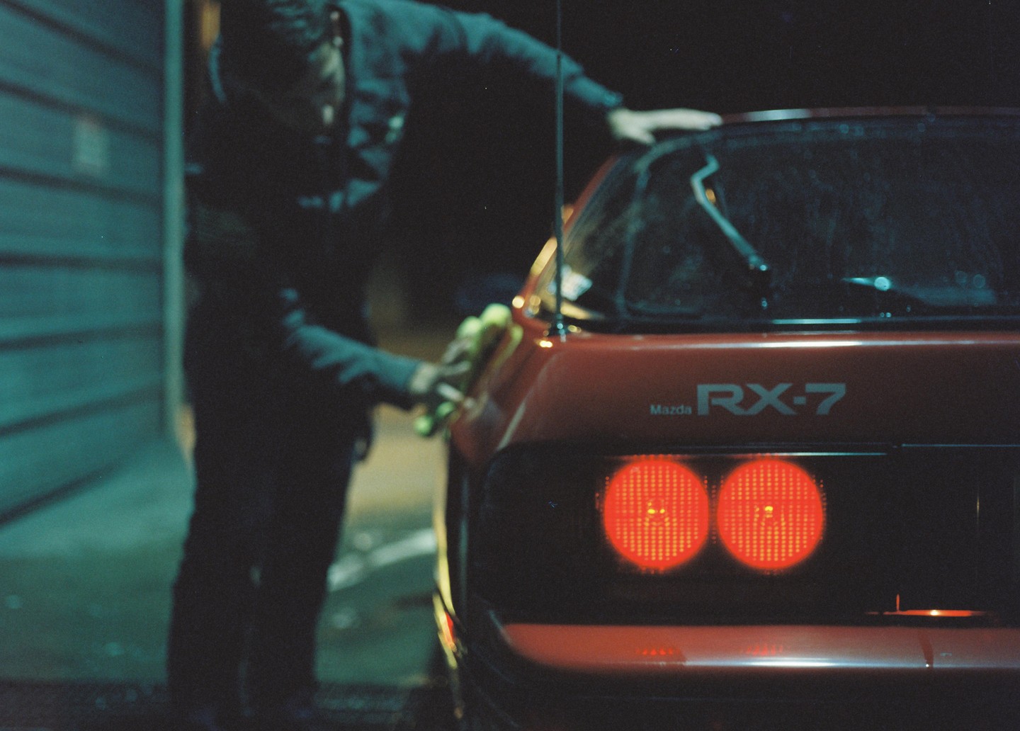 A person washes a red Mazda RX-7 at night, with the car's taillights glowing brightly.
