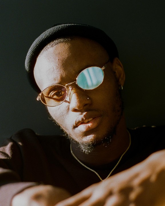 A man wearing a beanie and round sunglasses looks directly at the viewer.