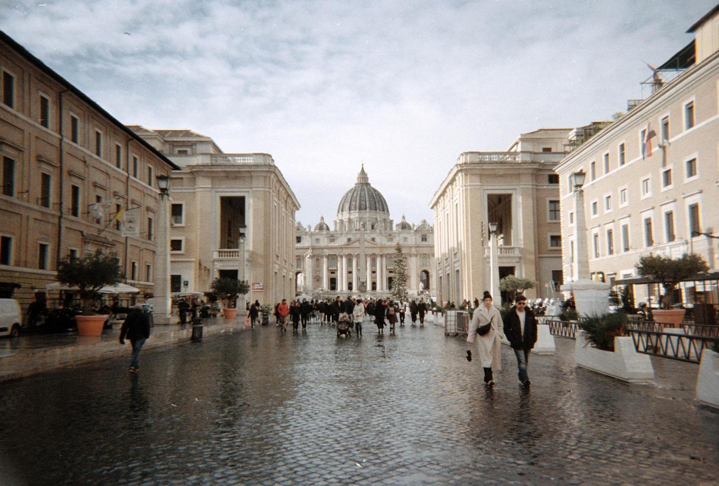 A wide view of St. Peter's Basilica with a Christmas tree in front, and people walking on a wet cobblestone street.