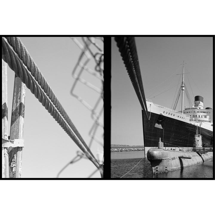 A large ship named "Queen Mary" and a submarine are docked in a harbor, viewed through a fence and cables.