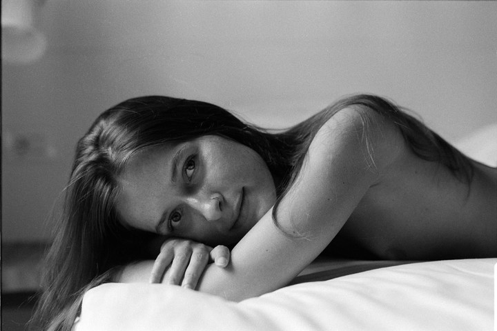 A woman with long hair is lying on a bed, looking towards the viewer.