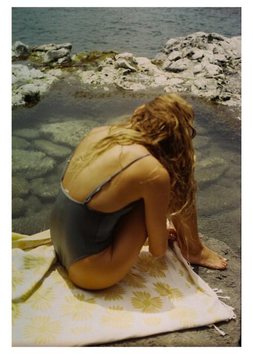 A woman in a swimsuit sits on a towel by the water's edge, with rocks and the ocean visible in the background.
