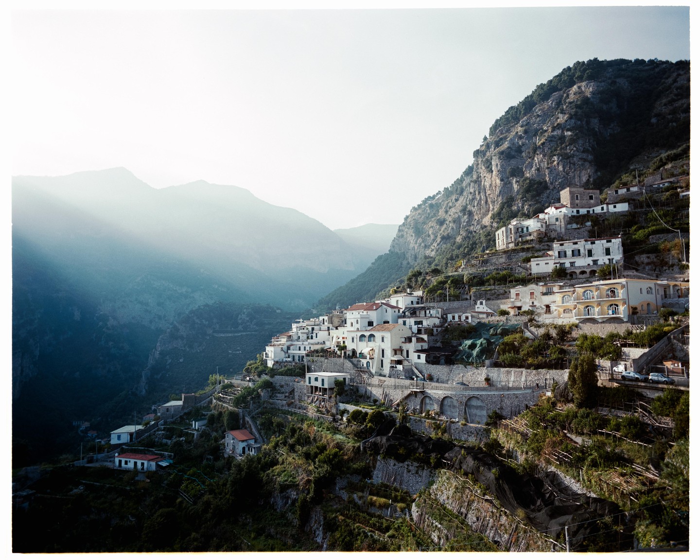 A village with white buildings is nestled into a steep, rocky mountainside with hazy mountains in the background.